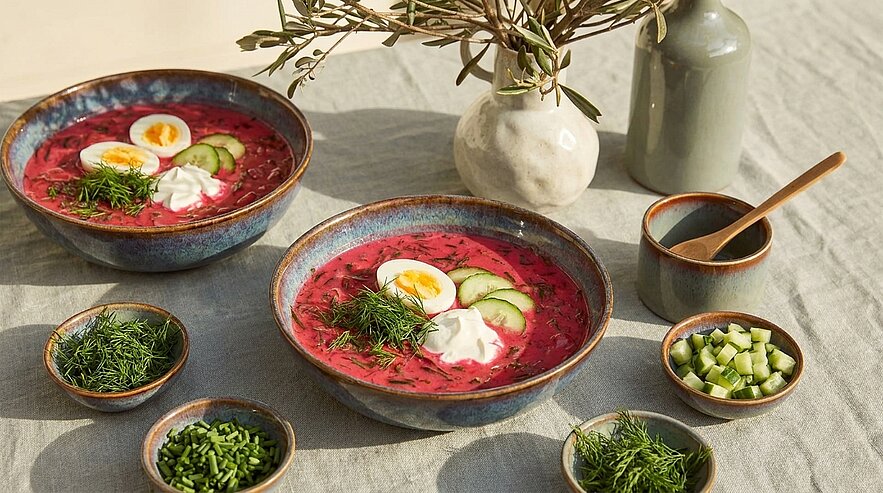Zwei Schalen kalte Rote-Bete-Suppe mit halbierten Eiern, Gurkenscheiben, einem Klecks saurer Sahne und frischem Dill, umgeben von kleinen Schälchen mit Kräutern und gewürfelter Gurke.