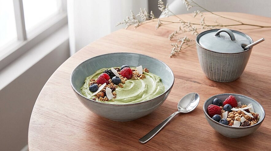 Eine große Schüssel mit cremigem, grünem Joghurt ist auf einem Holztisch plaziert, garniert mit Himbeeren, Heidelbeeren und Granola. Nebenan steht eine kleine Schüssel mit Himbeeren und Heidelbeeren. Ein Teelöffel liegt daneben, und im Hintergrund ist ein kleiner Behälter sichtbar.