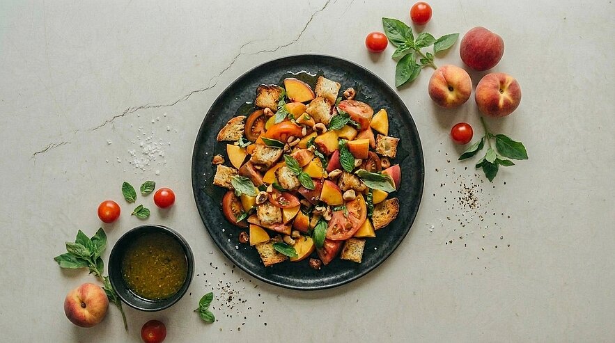 Teller mit gegrillten Pfirsichstücken, Cherry‑Tomaten und frischem Basilikum, beträufelt mit Vinaigrette; daneben eine Schale Dressing und einige ganze Pfirsiche.