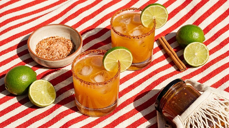 Zwei orangefarbene Cocktails mit Eis und Limettenscheiben, Glasränder mit Gewürzsalz, daneben Limetten und eine Schale mit Gewürzen.