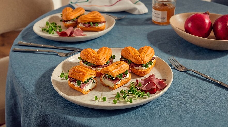 Teller mit vier herzförmigen Blätterteig-Sandwiches, gefüllt mit Frischkäse, Räucherlachs und Sprossen; daneben einige Scheiben Rohschinken.