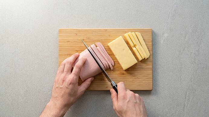 Hände schneiden Wurst mit Messer in dünne Scheiben auf einem Holzbrett; daneben gestapelte Käsescheiben.