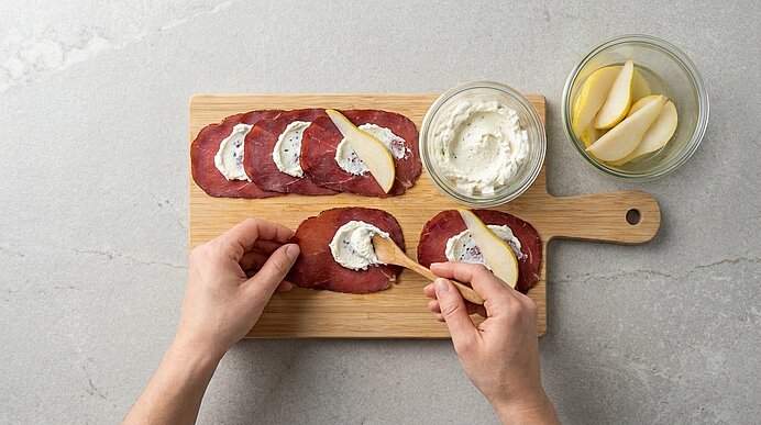 Zwei Hände streichen Frischkäsecreme mit einem Holzlöffel auf Bündnerfleisch-Scheiben auf einem Holzbrett.