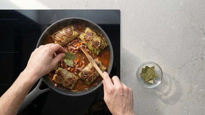 Zwei Hände rühren in einem Topf auf dem Herd gefüllte Kohlrouladen in Tomatensauce; eine Hand setzt ein Lorbeerblatt dazu, neben dem Topf liegt eine Schale mit weiteren Lorbeerblättern.