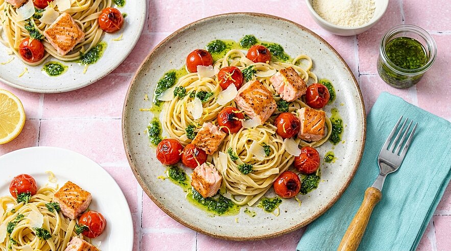 Teller mit Spaghetti‑Nestern, gebratenen Lachsfiletstücken, gerösteten Cherrytomaten, grüner Pesto‑Soße und Parmesanspänen.