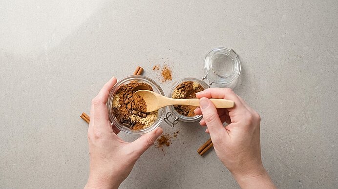 Zwei Hände füllen mit einem Holzlöffel ein kleines Glasgefäß mit gemahlenem Zimt; Zimtstangen liegen daneben.