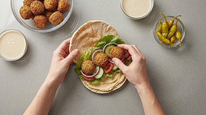 Zwei Hände legen knusprige Falafel in ein mit Hummus, Salat und Gemüse belegtes Brötchen.