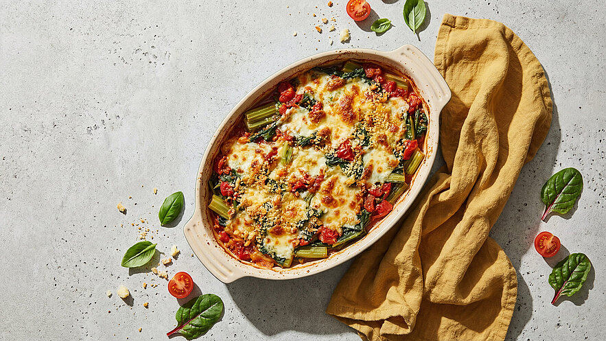 Ein Auflauf in einer ovalen, hellen Keramikform, gefüllt mit Spinat, Tomaten und Stangensellerie, überbacken mit geschmolzenem Käse. Um die Form liegen einige frische Tomaten, Basilikum- und Spinatblätter sowie ein gelbes Küchentuch.