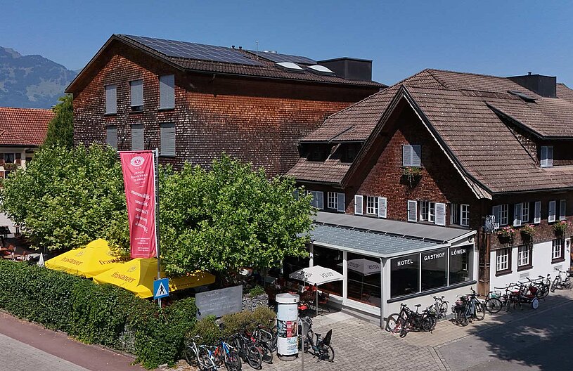 Ein zweigeschossiges Gebäude mit Holzfassade und Satteldach beherbergt das Hotel Gasthof Löwen. Vor dem Gebäude stehen Fahrräder und Tischbereiche im Freien. Bäume und Sonnenschirme verleihen der Umgebung eine einladende Atmosphäre. Im Hintergrund sind Berge sichtbar.