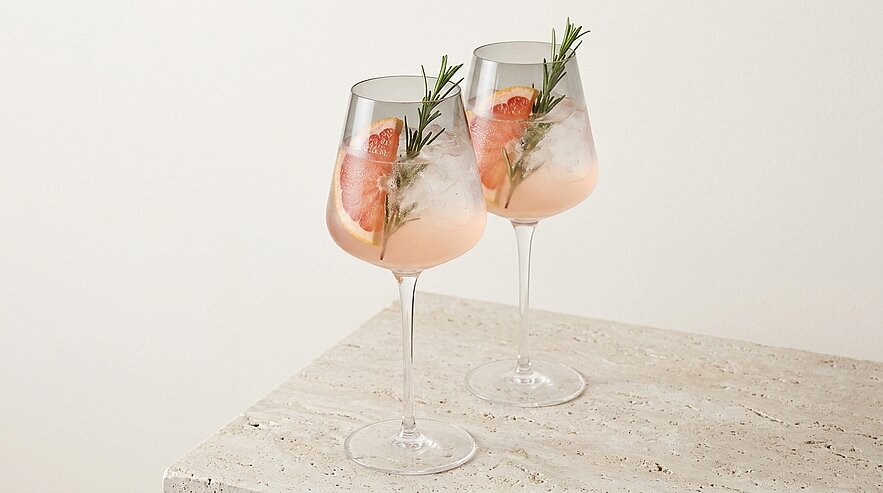 Zwei elegante Weingläser mit rosa Cocktail, Eiswürfeln, Grapefruitscheiben und Rosmarinzweigen auf hellem Steinuntergrund.