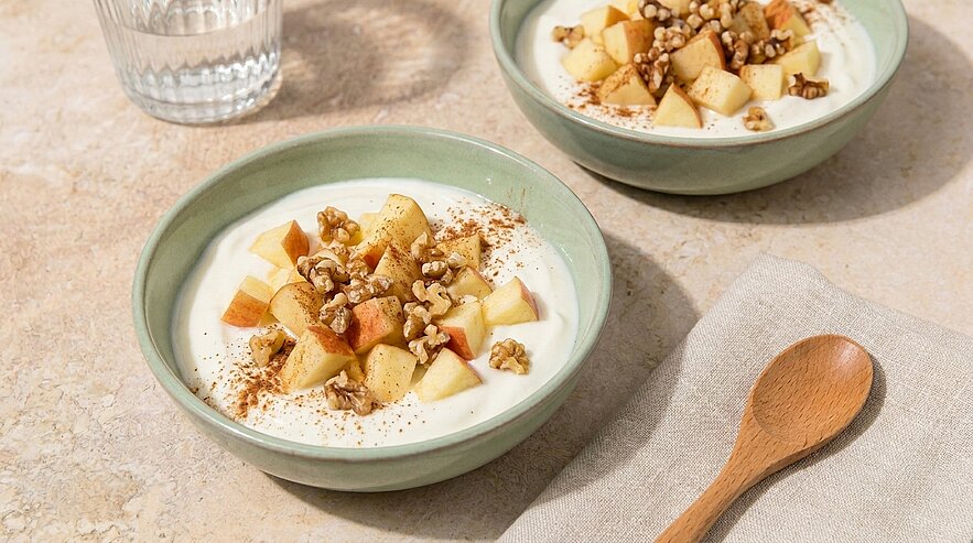 Zwei grüne Schalen mit Joghurt, gewürfelten Äpfeln, Walnussstücken und Zimt; daneben ein Holzlöffel.