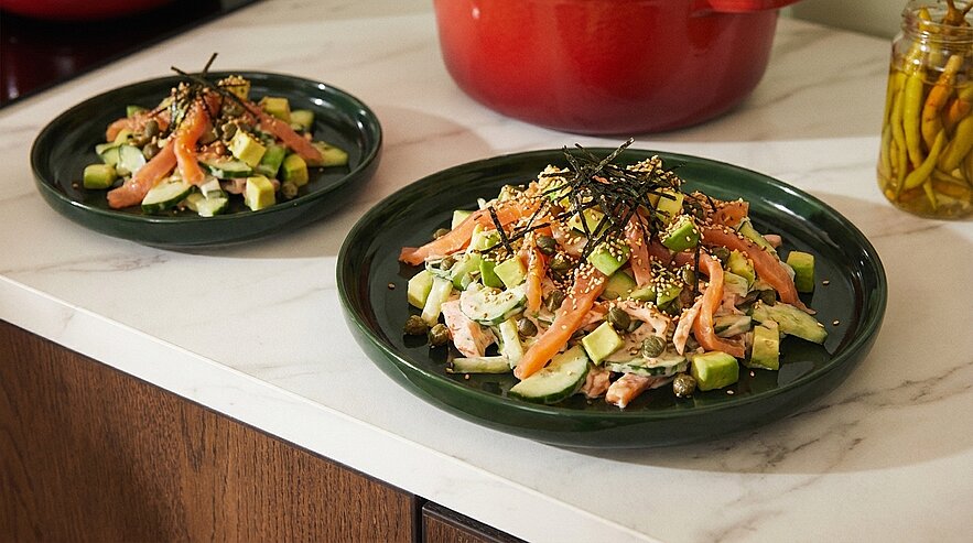 Zwei grüne Teller mit Salat aus Räucherlachsstreifen, Avocadowürfeln, Gurke, Frühlingszwiebeln, Sesam und dünnen Nori-Algenstreifen.
