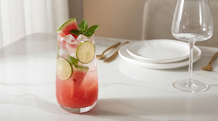 Glas mit rosafarbenem Wassermelonen-Cocktail auf Eis, dekoriert mit Limettenscheiben, Melonenkeil und Minzblatt.