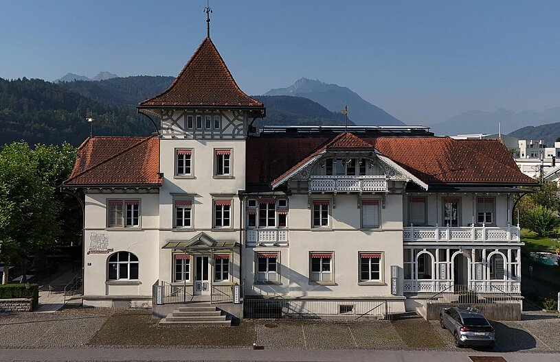 Das Bild zeigt ein historisches Gebäude mit einem markanten, spitzen Dach und einem Erker. Es hat helle Fassaden mit mehreren Fenstern und Verzierungen. Vor dem Gebäude sind Pflanzen und ein Parkplatz zu sehen, vor dem sich ein graues Auto befindet. Im Hintergrund erheben sich Berge.
