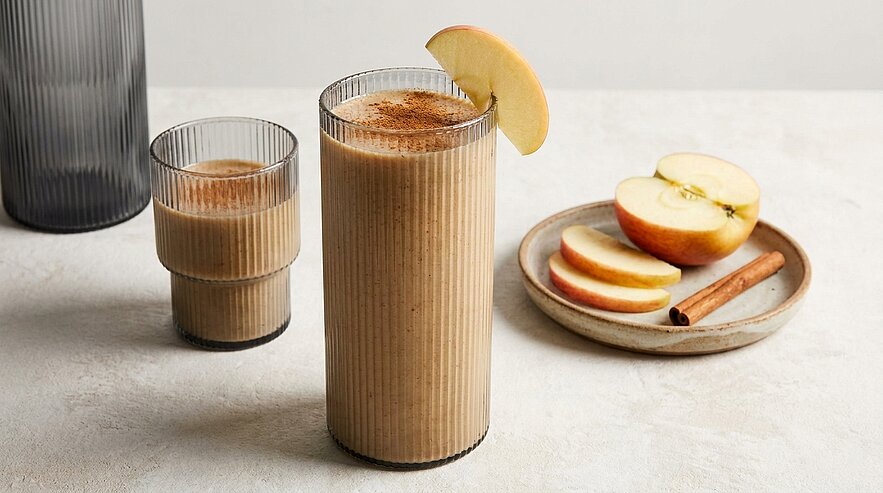 Auf der Abbildung sind zwei Gläser mit einem cremigen Apfel-Smoothie zu sehen, der mit einer Apfelscheibe und Zimt dekoriert ist. Daneben liegt ein Teller mit geschnittenen Äpfeln und einer Zimtstange. Die Gesamtästonung vermittelt einen frischen, gesunden Genuss.