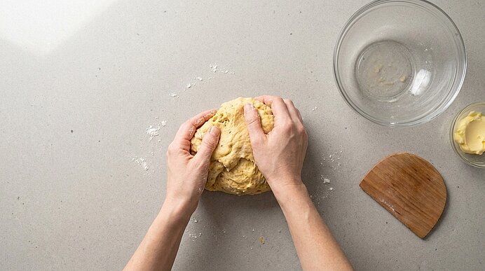 Zwei Hände kneten einen Teig auf einer Arbeitsfläche; daneben eine leere Rührschüssel, ein Teigschaber und etwas Butter.
