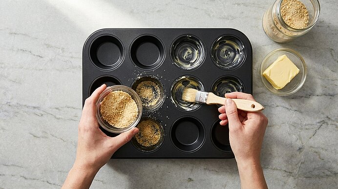 Zwei Hände bestreichen eine Muffinbackform mit geschmolzener Butter und drücken Keksbrösel als Boden in mehrere Mulden; neben der Form sind Butterstück und Schale mit Bröseln zu sehen.