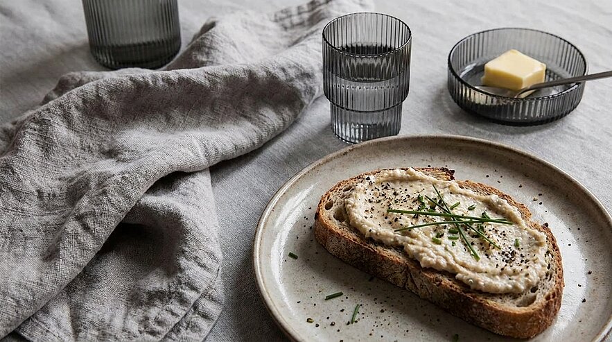 Scheibe rustikales Brot auf einem Teller, bestrichen mit cremigem Aufstrich und mit frischen Kräutern garniert; kleine Schale mit Butter daneben.