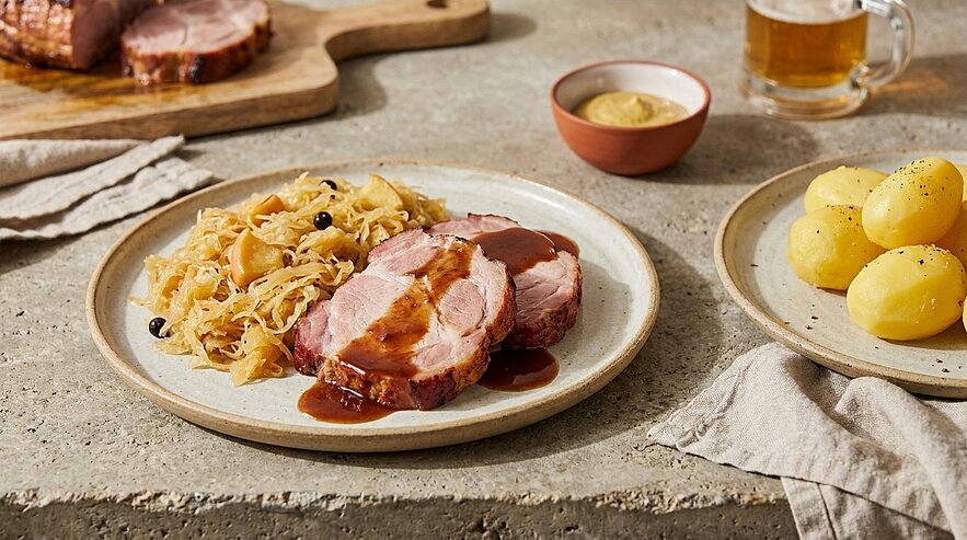 Teller mit zwei Scheiben Schweinebraten in dunkler Bratensoße, dazu Sauerkraut; daneben ein Teller mit Salzkartoffeln.