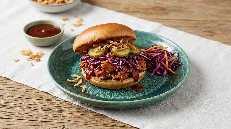 Pulled-Pork-Burger mit Barbecue-Soße, Gewürzgurken und knusprigen Röstzwiebeln, dazu Rotkohlsalat auf einem grünen Teller.