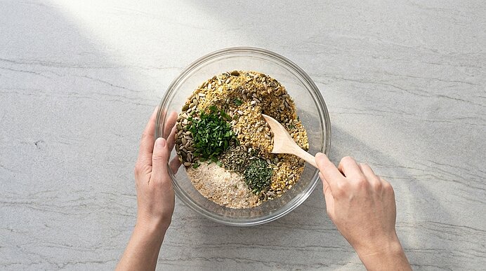 Zwei Hände rühren mit einem Holzlöffel in einer Glasschüssel eine Mischung aus Haferflocken, Samen und gehackten Kräutern.