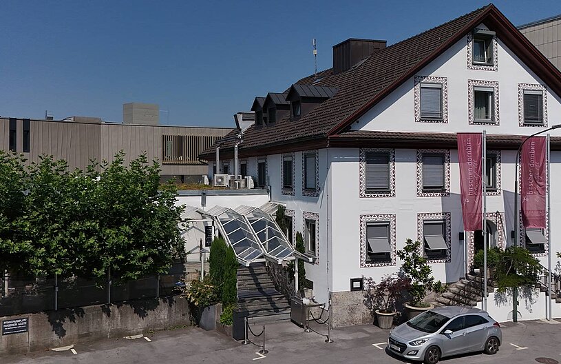 Ein helles, dreigeschossiges Gebäude mit einem giebelbegrünten Dach und dekorativen Fassadenelementen. Vor dem Eingang befindet sich eine Treppe mit einem Glasdach. Um das Gebäude sind Bäume und parkende Autos zu sehen. Im Hintergrund steht ein moderner, schlicht gestalteter Gebäudekomplex.