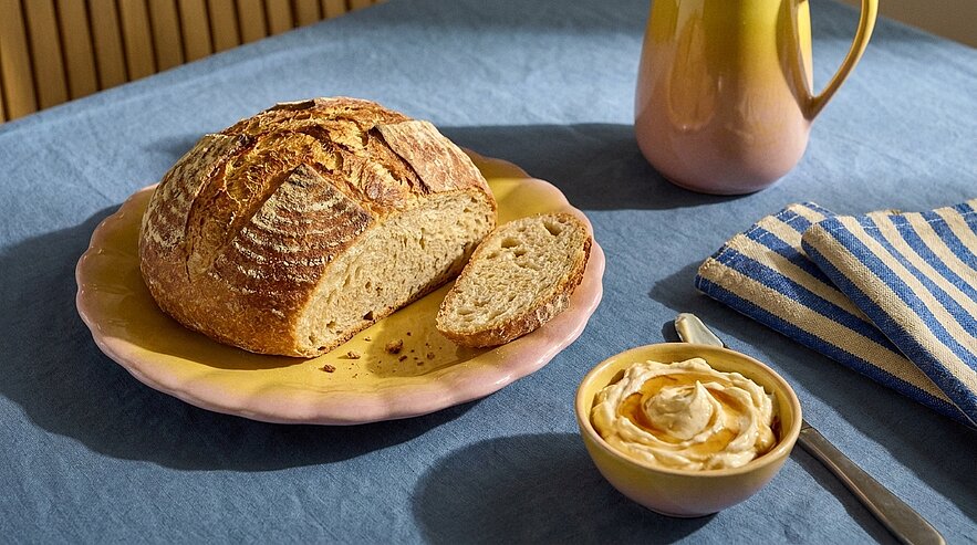 Ein frisch gebackenes rundes Brot auf einem gelben Teller, ein Stück ist davon abgeschnitten. Neben dem Brot befindet sich eine Schale mit cremiger Butter und ein Messerset. Im Hintergrund liegt ein gestreiftes Tuch.