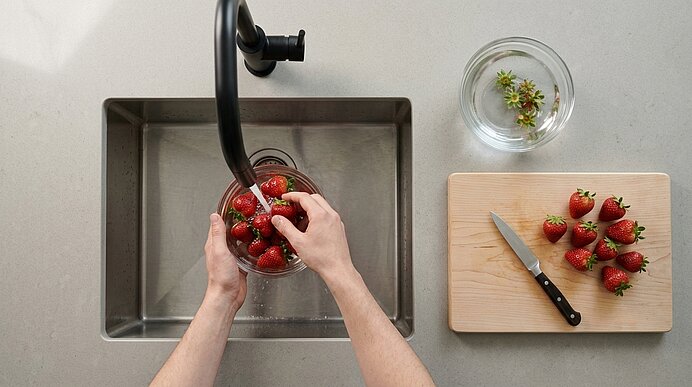 Zwei Hände waschen Erdbeeren im Spülbecken unter Wasser. Daneben liegen Messer, Holzbrett und Erdbeerstrünke.