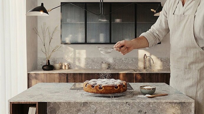 Teilweise sichtbare Person in Schürze bestäubt einen runden, frisch gebackenen Kuchen auf einem Gitter mit Puderzucker in einer modernen Küche.