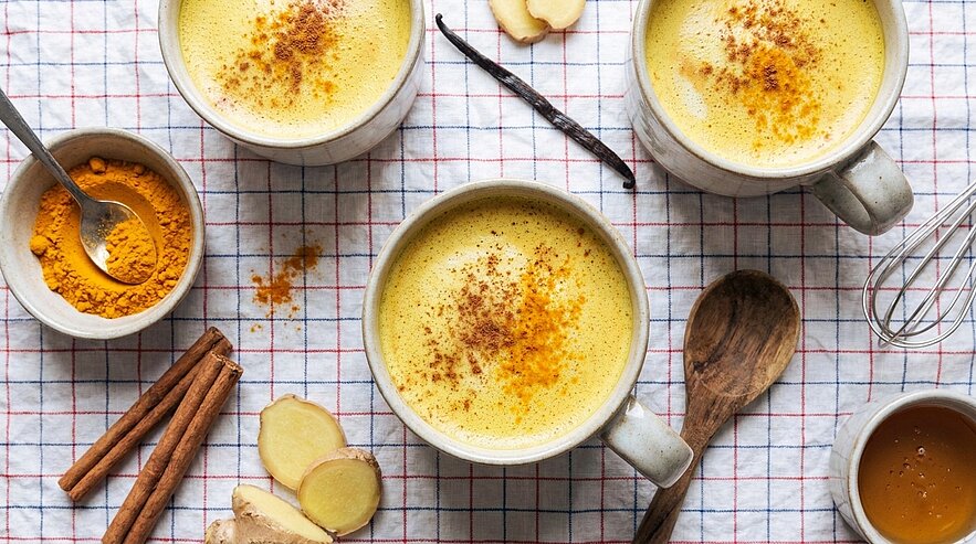 Drei Tassen goldene Milch (Kurkuma-Latte) mit Gewürzstreuseln auf kariertem Tuch, umgeben von Kurkumapulver, Zimtstangen, Ingwerscheiben und Honig.