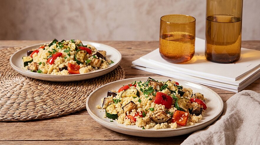 Zwei Teller mit Couscous-Salat, vermischt mit gerösteten Auberginen, Paprika, Kirschtomaten und frischen Kräutern, auf einem rustikalen Holztisch, daneben zwei bernsteinfarbene Trinkgläser.