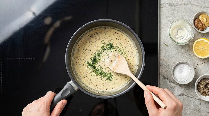 Hände rühren eine cremige Kräutersauce in einem Topf mit einem Holzlöffel; die Oberfläche ist mit frisch gehacktem Dill bestreut.