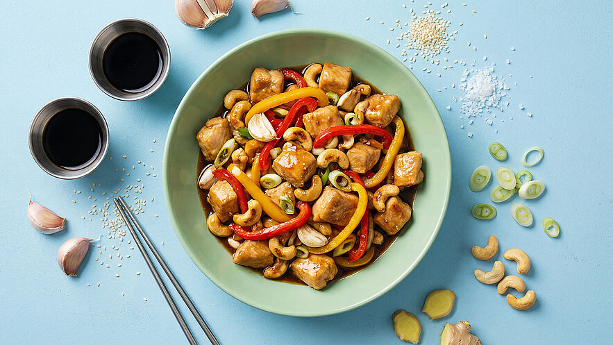 Eine grüne Schüssel enthält würfelig geschnittenes Hähnchenfleisch, bunten Paprika, Cashewkerne und Knoblauch in einer dunklen Sauce. Daneben liegen geriebener Ingwer, Frischgemüse und zwei kleine Schalen mit Sojasauce auf einem hellblauen Untergrund. Essstäbchen sind ebenfalls abgebildet.