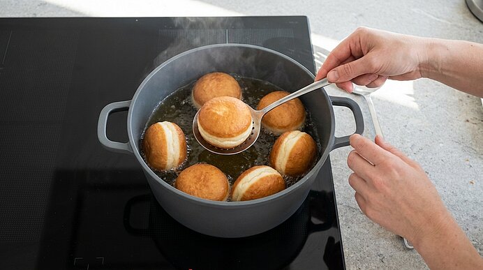 Hände wenden mit einem Löffel mehrere Krapfen (Berliner), die in heißem Öl in einem Topf auf dem Herd frittiert werden.
