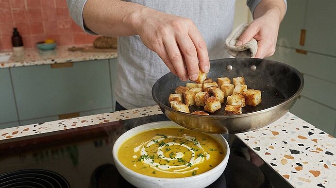 Hände kippen eine Pfanne mit goldbraunen Croutons in eine Schüssel cremiger Suppe, garniert mit einer Sahnespirale und gehackten Kräutern.