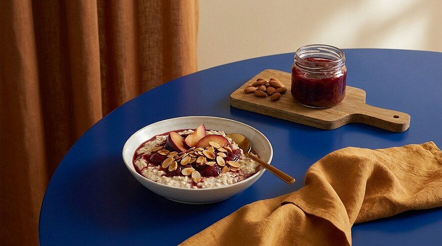 Schale Haferbrei mit Pflaumenstücken, Fruchtmus und Mandelblättchen, ein Löffel darin; daneben ein Glas Pflaumenmarmelade auf einem Holzbrett und ein gelbes Tuch auf blauem Tisch.