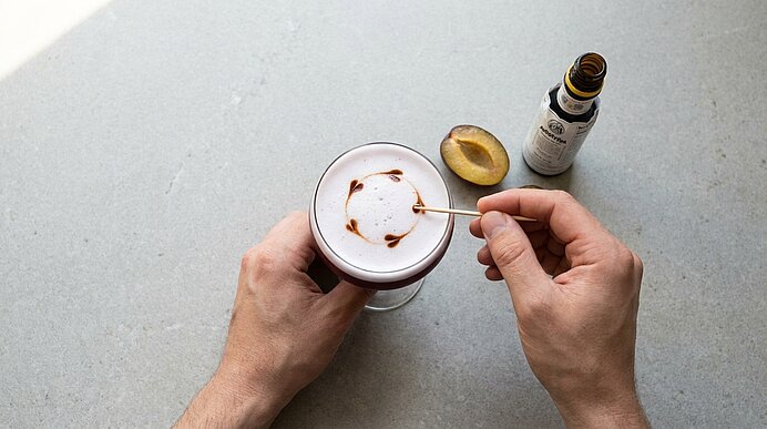 Zwei Hände halten eine Tasse Cappuccino; mit einem Zahnstocher wird ein Muster in den Milchschaum gezogen. Daneben liegt eine halbierte Pflaume und eine kleine Flasche.