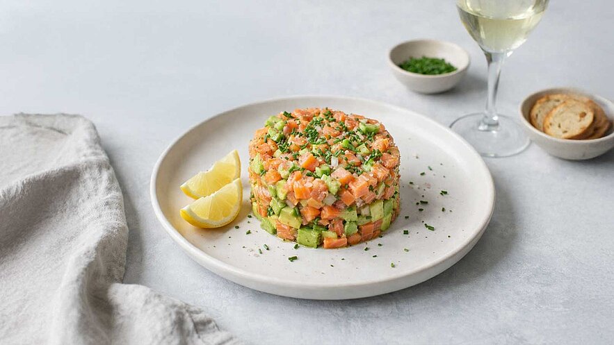Auf dem Bild ist ein Teller mit einem bunten Lachstatar zu sehen, das aus Lachs und Avocado besteht. Es ist mit frischen Kräutern garniert und drei Zitronenschnitzen dekoriert. Neben dem Teller steht ein Glas Weißwein.