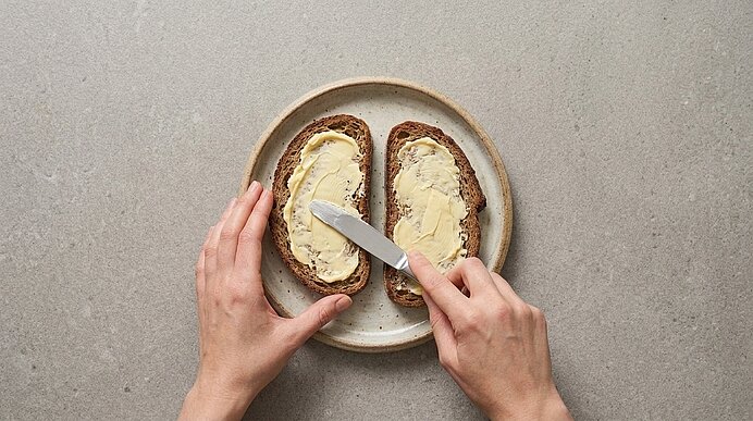 Zwei Hände bestreichen zwei Brotscheiben auf einem Teller mit einem Messer mit Butter.