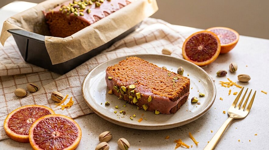 Scheibe saftiger Blutorangen‑Rührkuchen mit rosafarbener Glasur und gehackten Pistazien auf einem Teller; im Hintergrund Kastenform mit restlichem Kuchen und aufgeschnittene Blutorangen.