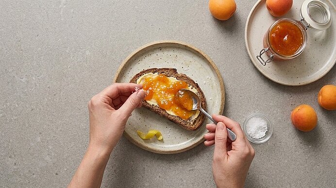 Zwei Hände verteilen Marillenmarmelade und Flockensalz auf einer mit Butter bestrichenen Scheibe Brot.