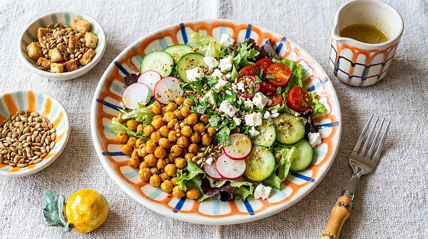 Bunter Teller auf gemusterter Keramik mit Salat: Blattsalate, Gurkenscheiben, Radieschen, Kirschtomaten, geröstete Kichererbsen und zerkrümelter Feta, bestreut mit Samen; ein kleines Gefäß mit Dressing daneben.
