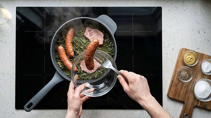 Zwei Hände geben Würstchen in eine Pfanne mit Grünkohl und Schinken auf einem Kochfeld; aus der Pfanne steigt Dampf.