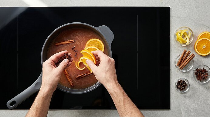 Zwei Hände legen Orangenscheiben und Gewürze (Zimtstangen, Sternanis, Nelken) in einen Topf mit heißem Punsch auf einem Glaskeramikherd; mehrere Schälchen mit Orangen und Gewürzen liegen daneben.