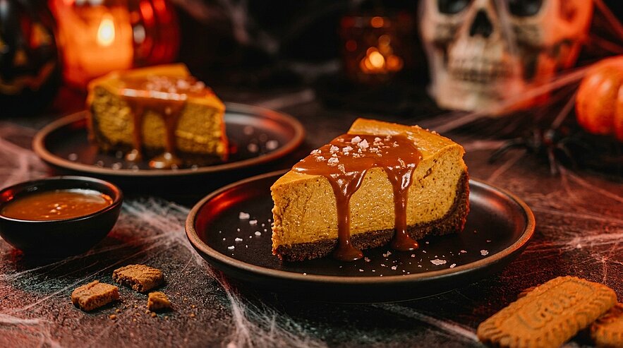 Nahaufnahme eines Stücks Kürbiskäsekuchen auf schwarzem Teller, mit Karamellsauce und Meersalz bestreut; im Hintergrund weiteres Stück, Schälchen Karamell und Gewürzkekse.
