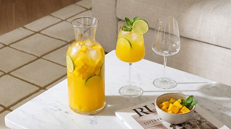 Glaskaraffe mit eisgekühltem gelben Fruchtsaft und Limettenscheiben, ein gefülltes Weinglas mit Limetten- und Minzgarnitur, ein leeres Weinglas und eine Schale mit Mangowürfeln auf einem weißen Marmor-Couchtisch.