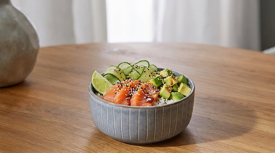Schüssel mit Poké-Bowl aus Reis, dünnen Lachsscheiben, Avocadowürfeln, Gurkenbändern, Sesam und einer Limettenspalte.