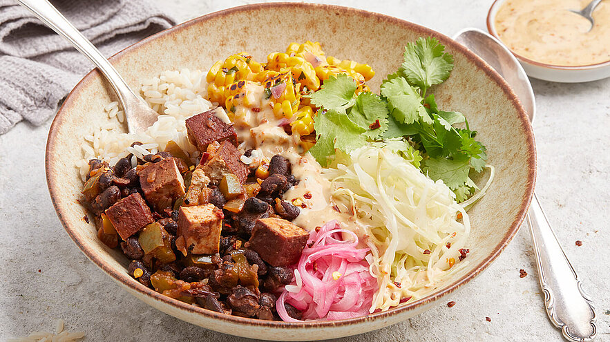 Eine Schüssel mit Reis, gebratenem Tofu, schwarzen Bohnen, süßer Mais, frischem Koriander, roten Zwiebeln und Krautsalat. Die Zutaten sind kunstvoll angeordnet und bieten eine bunte, abwechslungsreiche Mahlzeit.