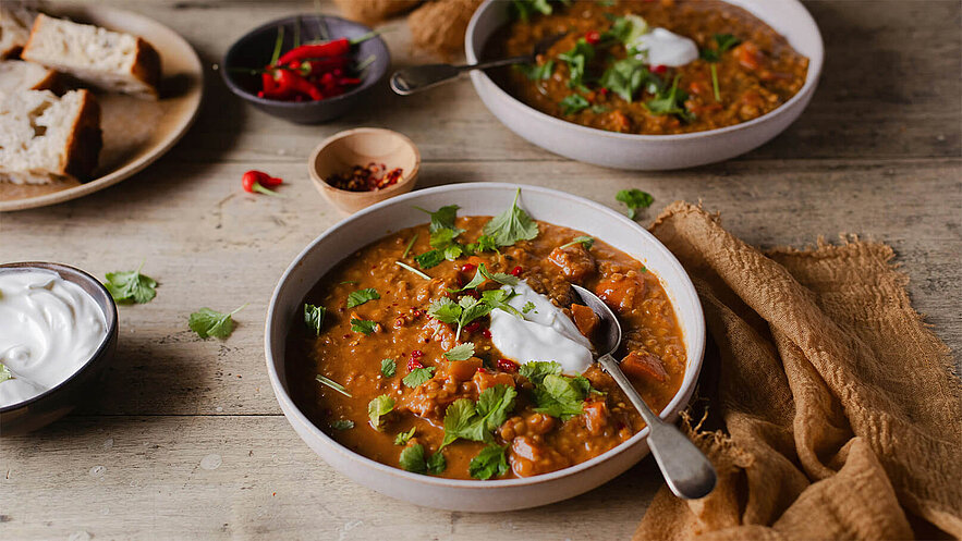 Zwei Schalen mit dampfender Suppe, garniert mit frischem Koriander und einem Klecks Joghurt, stehen auf einem Holztisch. Daneben sind Schalen mit Gewürzen und einige Stücke Brot zu sehen.