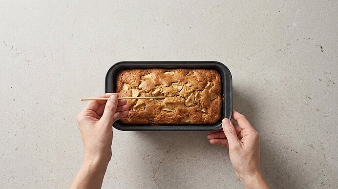 Zwei Hände halten eine Kastenform mit frisch gebackenem Bananenbrot; eine Hand führt einen Holzspieß zur Garprobe in das Brot.